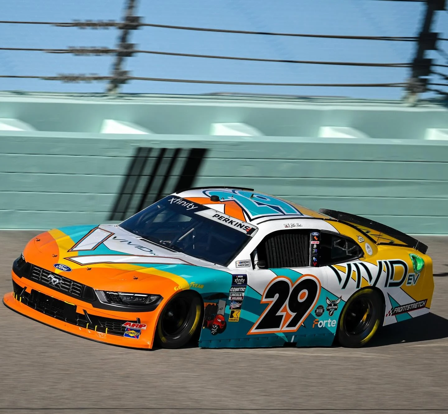 Orange, teal, white and yellow No. 29 Vivid EV Ford Mustang at speed against the Homestead-Miami wall